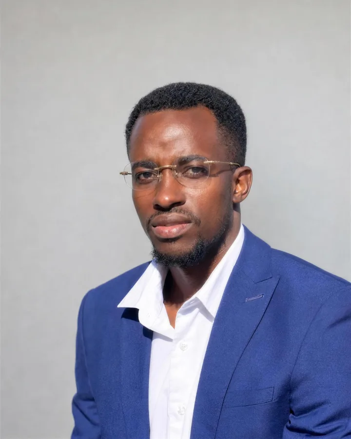 Jovan Baganja in a navy blazer and white shirt, wearing rimless glasses, against a plain grey wall.