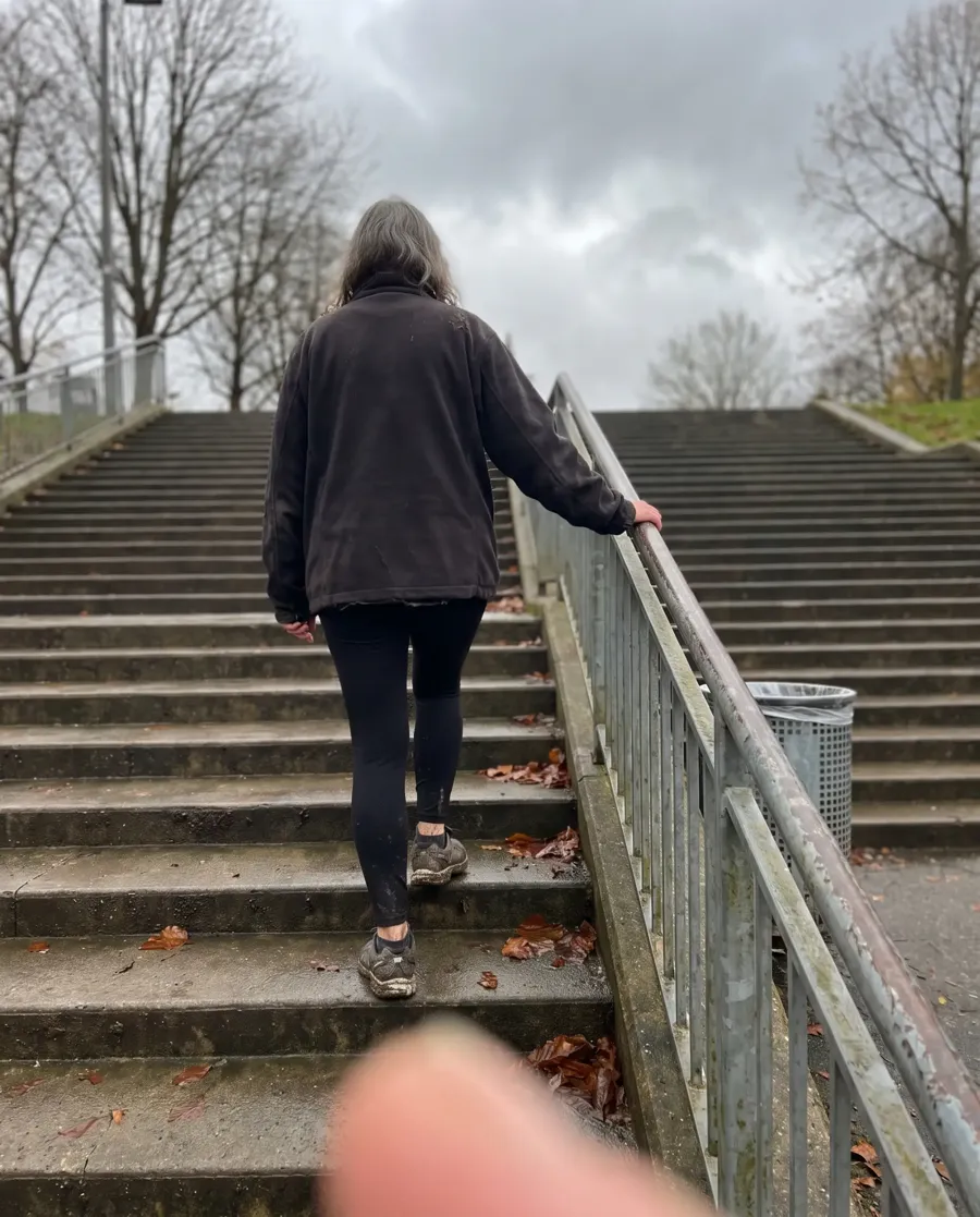 Candid phone photo from behind of a woman with grey hair in a fleece and leggings climbing outdoor concrete stairs on an overcast autumn day, a blurred thumb in the corner of the frame.